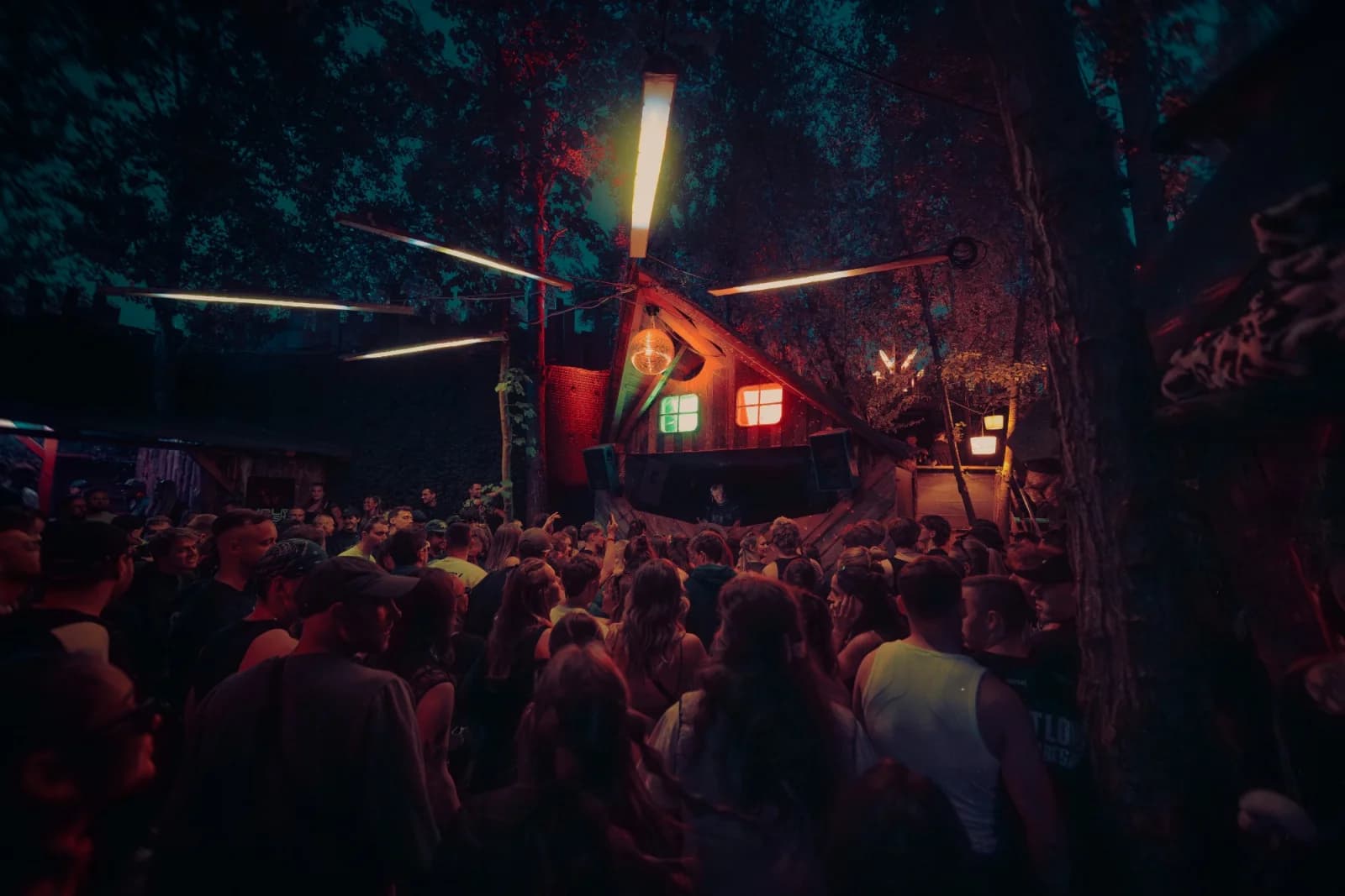 Westhafen Leipzig bei Nacht: Publikum vor einem DJ-Häuschen aus Holz zwischen Bäumen, mit Discokugel und bunten Fenstern